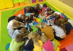 Grupa dzieci trzymająca się za dłonie siedzi pochylona w okręgu na chuście animacyjnej.
